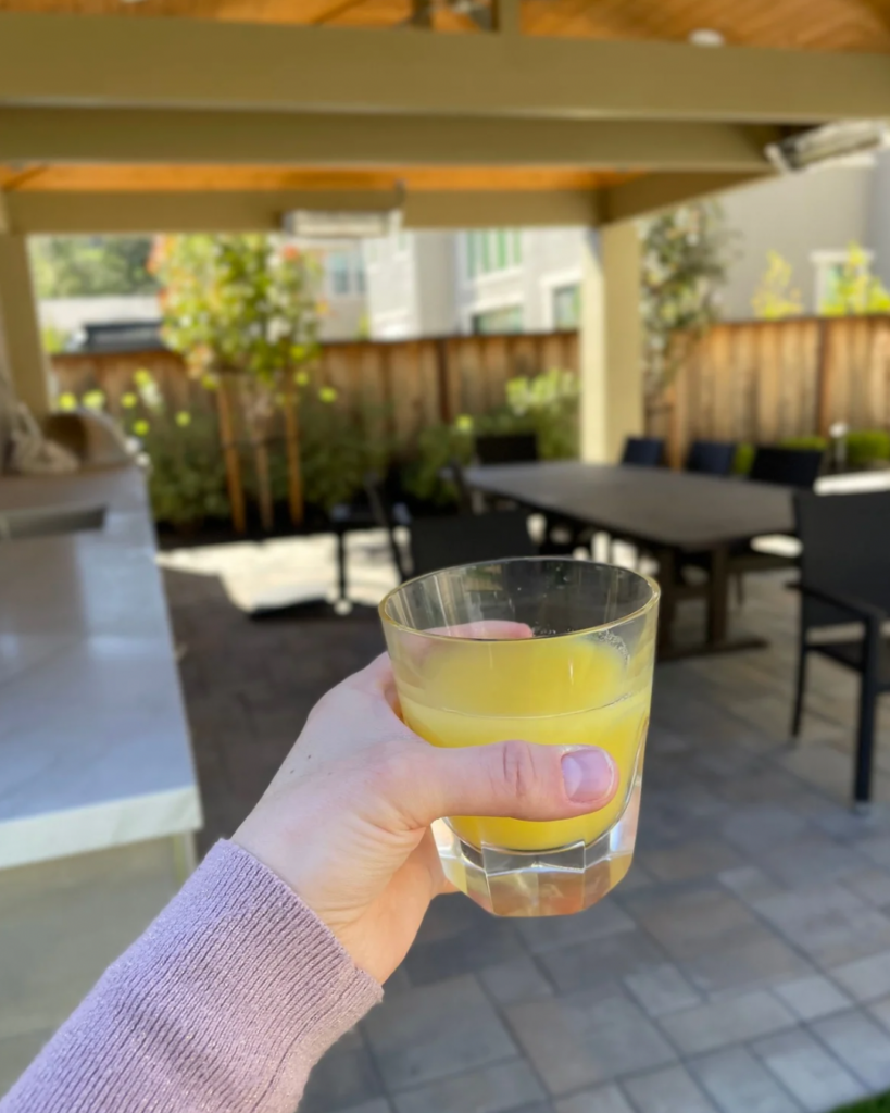 A hand holding a glass of orange juice in a covered outdoor patio area with a wooden table and chairs in the background.