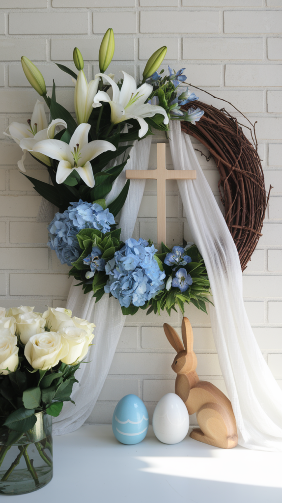 An Easter wreath with white lilies, blue hydrangeas, and a wooden cross, draped with white fabric, next to a wooden rabbit figurine and two decorative eggs, placed on a white surface against a brick wall.