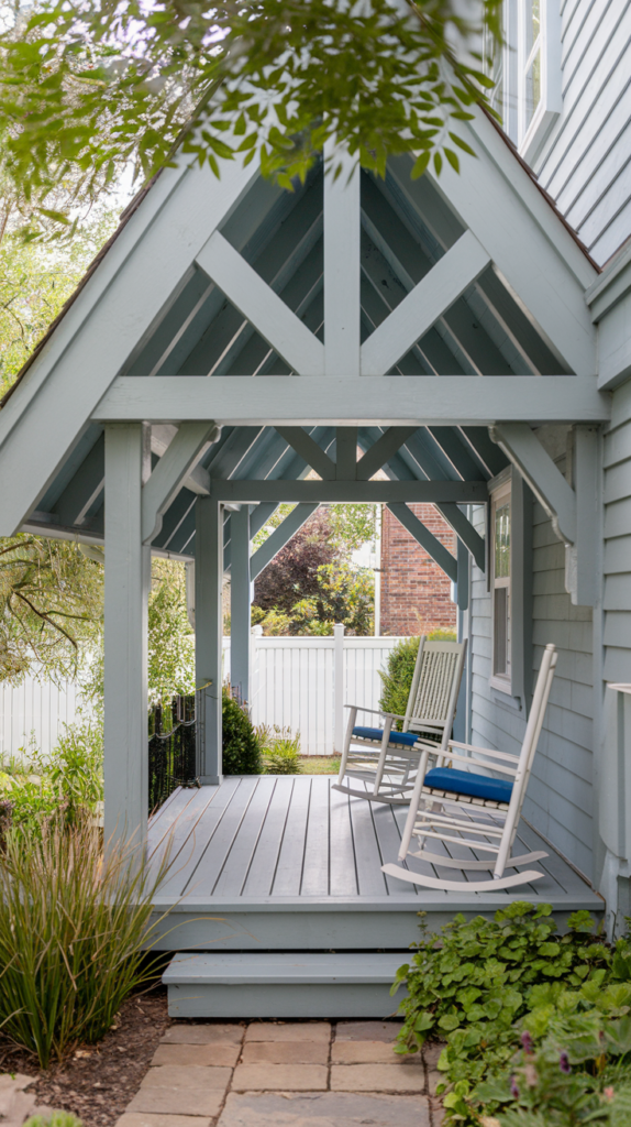 A quaint porch with light blue wooden beams and two white rocking chairs with blue cushions. The porch is surrounded by greenery and a white fence is visible in the background.