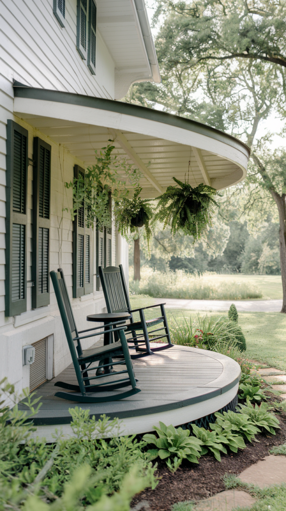 A curved porch featuring two dark green rocking chairs and a small table, with hanging ferns and a lush garden in the background.